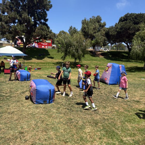 Kids playing in a park with inflatable barriers and trees in the background.