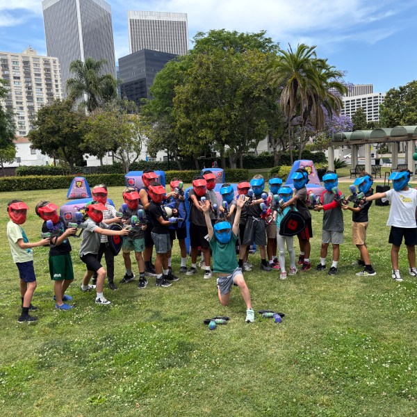 Children wearing helmets and holding blasters at an outdoor activity in a park with tall buildings.
